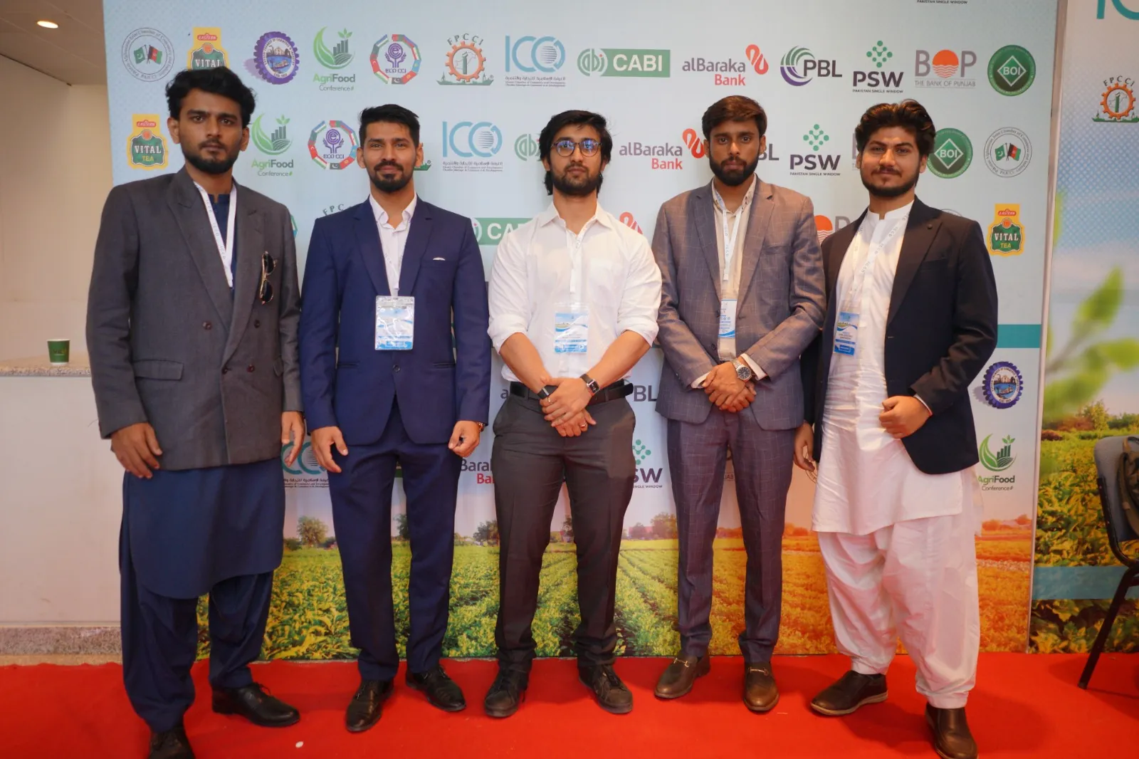 Five men in business and traditional dress standing together on a red carpet at an agriculture expo
