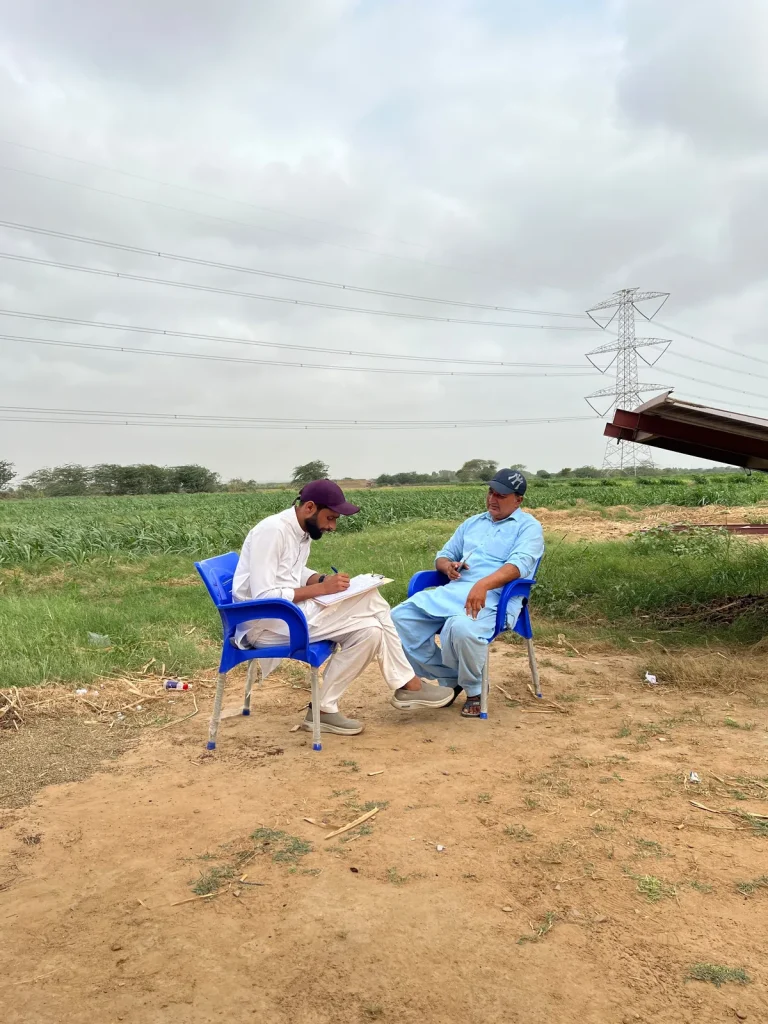 Two men seated on blue chairs on a dirt track beside a green field, one taking notes during the field visit