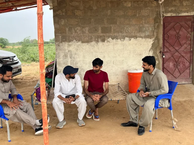 Men seated on chairs under a shelter beside a mud-brick wall during the field visit
