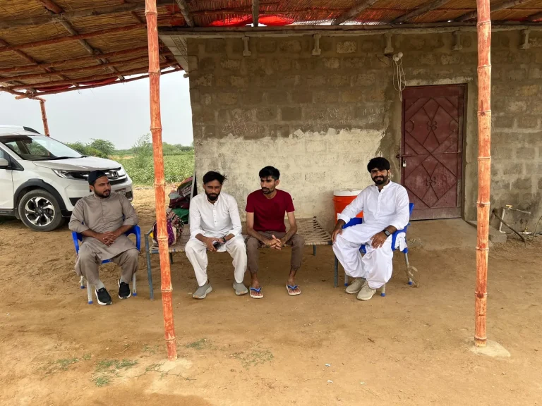 Men seated under a shelter next to a parked vehicle, with orange support poles